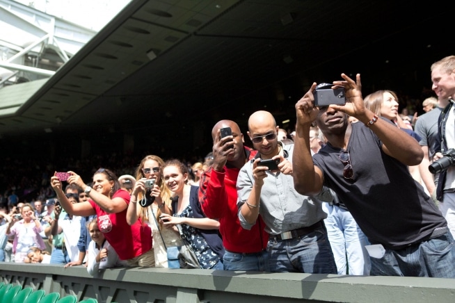 Spectators lifting phones and cameras beside a tennis court at Wimbledon.