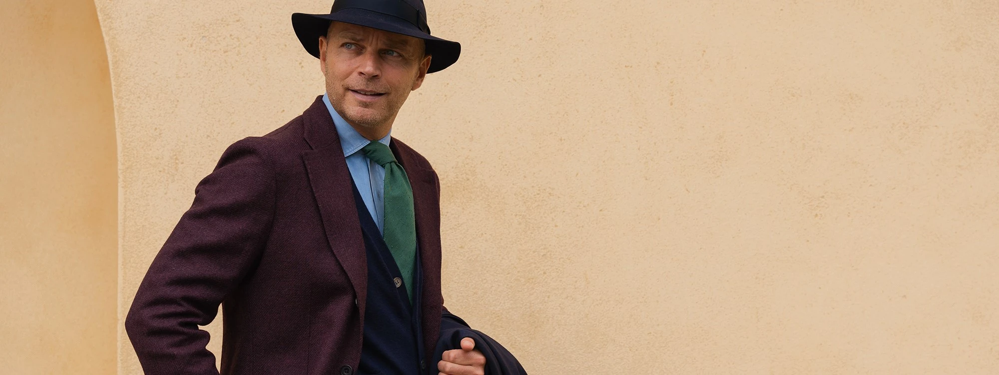 A man in a burgundy Rubinacci jacket and dark hat against a warm stone wall.