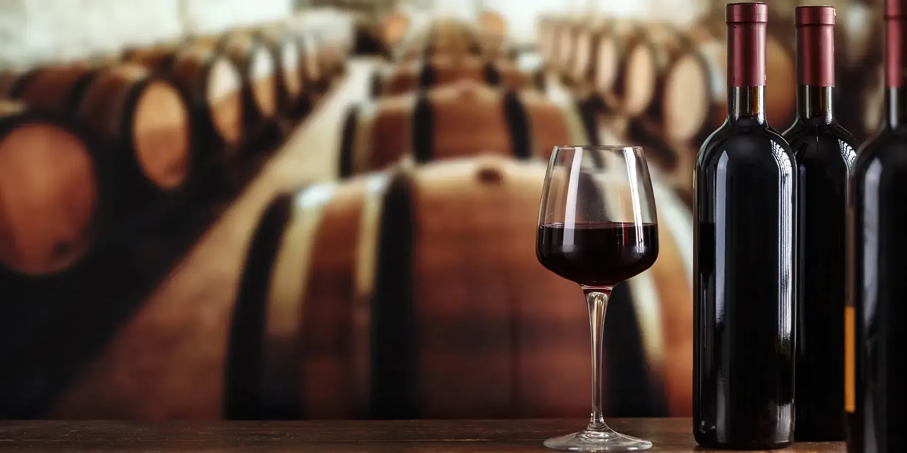 A glass of red wine beside dark bottles, with oak barrels in the cellar behind them.